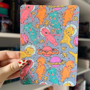 May include: A repeating pattern of colorful cartoon aliens with large eyes and antennas, wearing space suits, against a blue background with pink, orange, and yellow planets and stars.