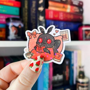 May include: A holographic sticker featuring a cartoon moth reading a book in bed with a cup of tea. The moth is wearing a red blanket and has red eyes. The sticker has a pink background with glitter and the text "Have you seen me?" on a small note.