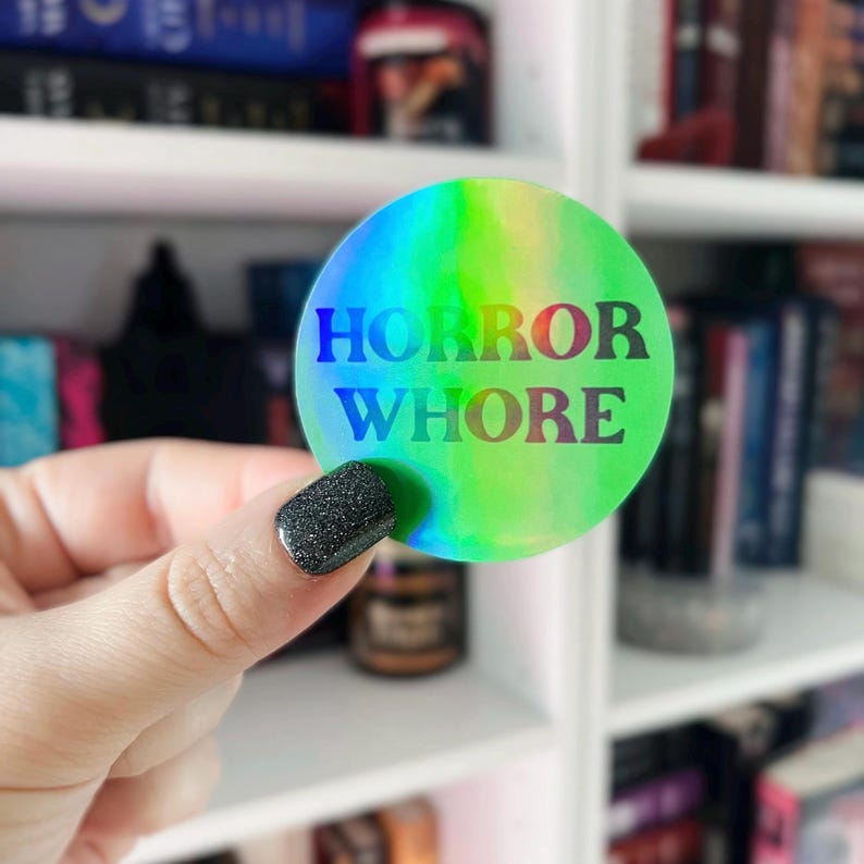 以下が含まれることがあります： 黒い文字で「HORROR WHORE」と書かれた、虹色の丸いステッカーです。