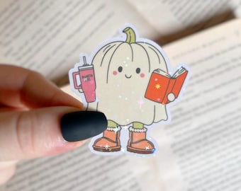 Puede incluir: Un personaje blanco con forma de calabaza que lleva botas marrones y sostiene un libro rojo y una taza rosa con el texto "Pumpkin Spice" en ella.