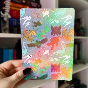 Peut inclure: Un carnet rectangulaire avec un design holographique coloré. La couverture présente un motif répété de créatures fantastiques, dont des dragons, des lapins et des fantômes, sur un fond étoilé. Les bords du carnet sont arrondis.