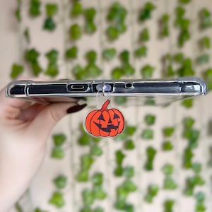 May include: Clear phone case with a dangling orange pumpkin charm. The pumpkin has a carved face with black details. The phone case is held against a background of green vines.