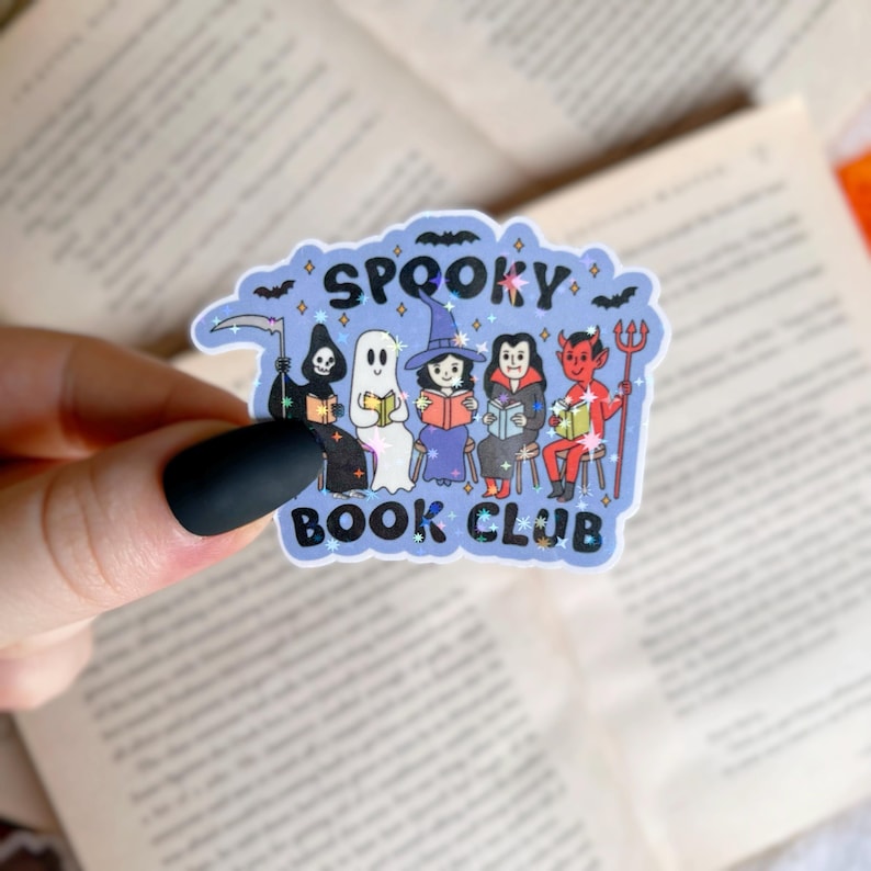 Op de afbeelding: Een blauwe en witte sticker met een glinsterende afwerking waarop een groep griezelige figuren te zien is die boeken lezen. De tekst luidt "Spooky Book Club".