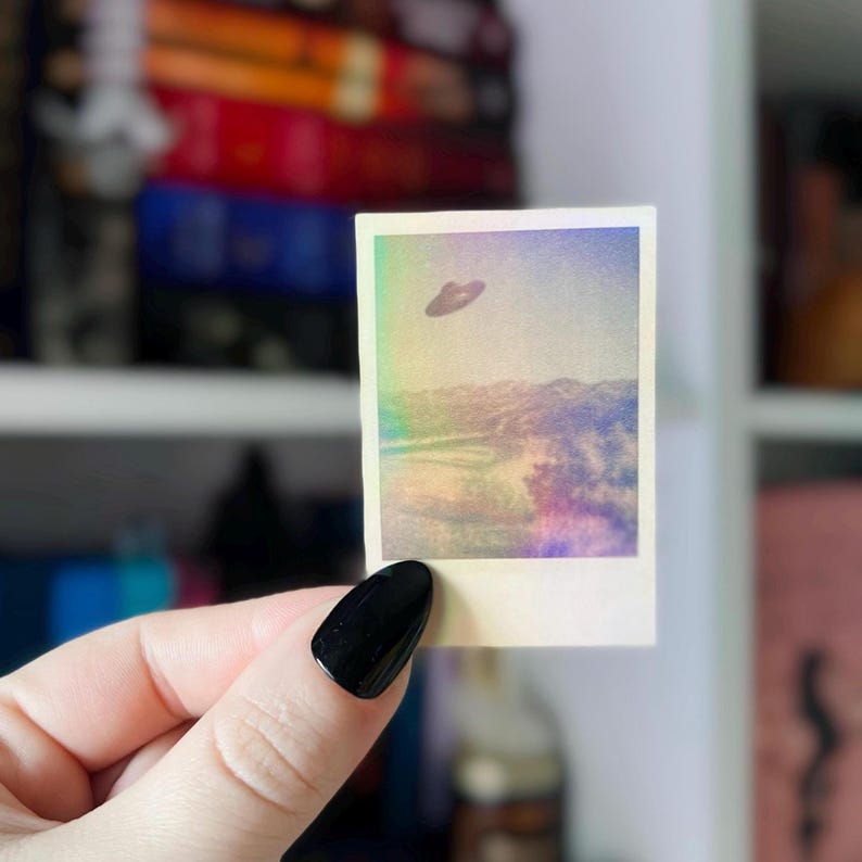 May include: A small, square photograph featuring a vintage-style image of a flying saucer hovering over a landscape. The image has a retro, faded color palette with a rainbow-like effect around the edges. The photograph is held by a hand with a black painted nail.