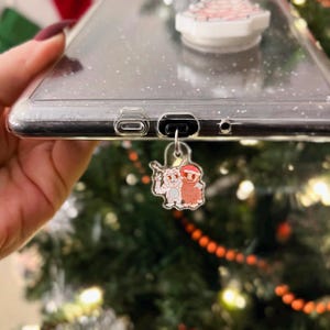 May include: A clear phone case with glitter and a cartoon charm. The charm features two cartoon characters, one white and one brown, wearing Santa hats. The phone case has a pop socket on the back.