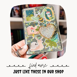 Puede incluir: Funda de tel&eacute;fono transparente decorada con pegatinas de una rana, una abeja, setas y un caracol. Un adorno en forma de coraz&oacute;n con purpurina iridiscente y ribete dorado est&aacute; centrado en la funda. El texto "find more JUST LIKE THESE IN OUR SHOP" est&aacute; en la parte inferior.