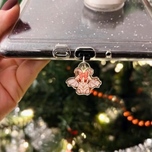 May include: A clear phone case with a glittery background and a pink and white cartoon character charm attached to the bottom. The charm is a small, winged creature with horns and a bow.