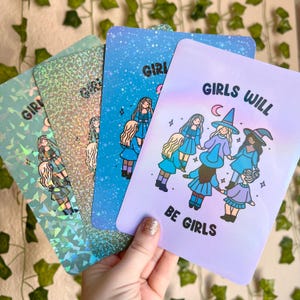 May include: Four colorful art prints featuring illustrations of girls. The prints have a variety of backgrounds, including holographic, blue with stars, and lavender with the text "GIRLS WILL BE GIRLS". The illustrations depict girls in dresses.