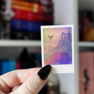 May include: A small, square print featuring a bridge with a winged creature in the sky. The image has a vintage, faded look with a color gradient of pink, purple, and green. The print is held by a hand with a black painted nail.