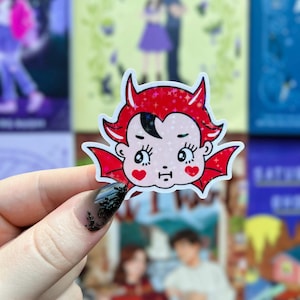 May include: A sticker featuring a cartoon devil with red horns, wings, and hair. The devil has a pale face with heart-shaped cheeks and a small mouth. The sticker has a holographic, glittery finish.
