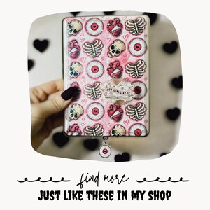 May include: A rectangular e-reader case with a pink background featuring a repeating pattern of skulls, eyeballs, rib cages, and hearts. A circular sticker on the front displays an open book with the text HOT GIRLS READ. A small circular charm hangs below.