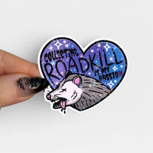 Op de afbeelding: Een hartvormige holografische sticker met een grijs en wit opossum met de tong uit de bek. De achtergrond is een donkerblauwe sterrenhemel met de tekst "COLLECTING ROADKILL IS MY PASSION" in dikke zwarte hoofdletters.