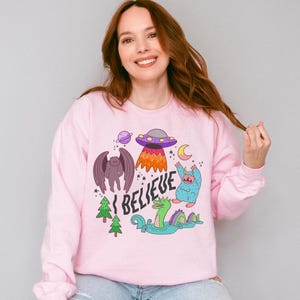 May include: A light pink long-sleeve sweatshirt featuring a colorful graphic print of a purple UFO, a planet, a crescent moon, a winged creature, a blue yeti, and a green sea monster in water, with the text I BELIEVE written in black letters.