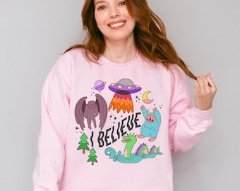 May include: A light pink long-sleeve sweatshirt featuring a colorful graphic print of a purple UFO, a planet, a crescent moon, a winged creature, a blue yeti, and a green sea monster in water, with the text I BELIEVE written in black letters.