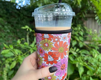May include: A reusable coffee cup sleeve with a colourful floral pattern featuring ghosts, pumpkins, and spiderwebs. The sleeve is made of neoprene and is designed to keep drinks cold or hot.