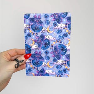May include: A rectangular notebook with a repeating pattern of cartoon bats, moons, clouds, and stars in shades of blue, purple, and yellow. The notebook is held by a hand with a red painted nail and a snake ring.