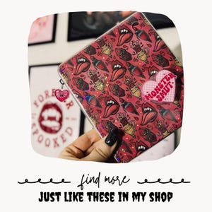 May include: A square zippered pouch with a red background featuring a repeating pattern of vampire mouths, fangs, and potion bottles. A pink heart-shaped sticker with the text MONSTER SMUT is attached to the front, and a small heart charm hangs from the zipper.