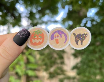 May include: Three round iridescent stickers with Halloween themes. The stickers feature a jack-o'-lantern, a ghost, and a black cat.
