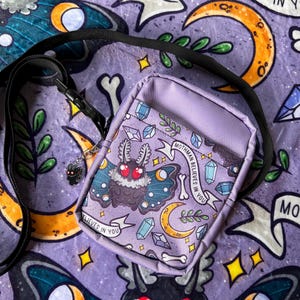 May include: A purple crossbody bag with a graphic print of a moth, crystals, and the text "Mothman Believes in You".