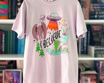 May include: A light purple short-sleeve t-shirt featuring a graphic print with the text I BELIEVE. The design includes a purple UFO, a brown moth-like creature, a blue yeti, a green sea serpent, a crescent moon, a planet, and green pine trees.