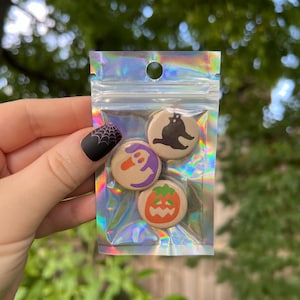 May include: A set of four Halloween-themed button badges. The badges are round and feature a black cat, a ghost, a pumpkin, and a spider web. The badges are packaged in a clear plastic bag with a holographic finish.