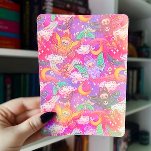 Puede incluir: Una tarjeta estampada con ilustraciones de criaturas aladas, lunas, arcoíris y nubes en una combinación de colores vibrantes. El fondo es una mezcla de rosa, morado y amarillo. La tarjeta la sostiene una persona con esmalte de uñas negro.