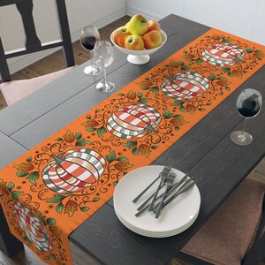 May include: Orange table runner with a repeating pattern of striped pumpkins, leaves, and vines. The runner is on a dark wood table with a white plate and silverware.