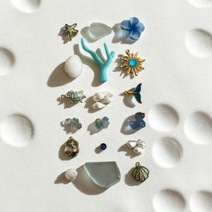 May include: An assortment of ocean-themed charms and decorative elements. Includes a starfish, seashells, a coral branch, a turtle, a sunburst, a duck, and other sea-inspired designs. The items are in various colors, including gold, blue, and white.