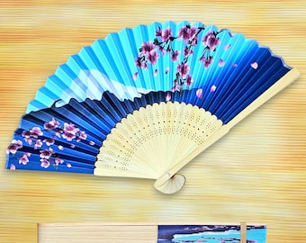 May include: A blue and white hand fan with a floral design. The fan features a light wood handle and a painted design of cherry blossoms and a mountain landscape. The fan is open, and a closed fan is visible below.