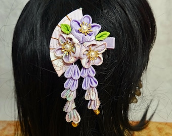 May include: Purple and white fabric flower hair clip with a fan design and dangling strands of fabric. The clip is worn in a woman's black hair.