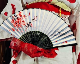 May include: A folding hand fan with a white background, featuring a painted design of red cherry blossoms on branches and small birds. The fan has a black frame with intricate cut-out details at the base and a pleated fabric surface.