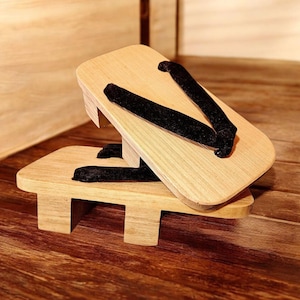 Puede incluir: Dos sandalias geta de madera con correas de tela negra. Las sandalias están hechas de madera de color claro y tienen una plataforma elevada. Las correas son de un material oscuro y aterciopelado. Las sandalias se exhiben sobre una superficie de madera.