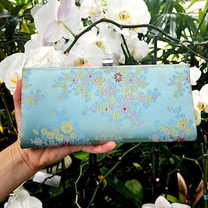 May include: A rectangular clutch with a light blue background and a floral pattern in yellow, pink, and blue. The clutch has a silver clasp and is held in a hand.