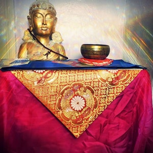May include: A golden statue of Buddha sitting on a red and gold patterned cloth with a singing bowl and cushion in front of it.