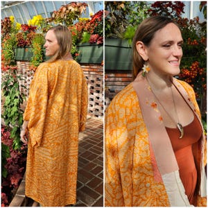 May include: A long, flowing orange kimono with a cream and orange floral pattern. The kimono has wide sleeves and a peach-colored collar with floral embroidery. The garment is worn over a rust-colored dress.