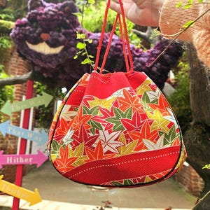 Puede incluir: Una bolsa con cordón de color rojo que presenta un patrón vibrante de hojas de arce en tonos naranja, amarillo, verde y blanco. El bolso tiene dos asas finas de cuerda roja y una base de tela roja sólida con un detalle de línea punteada blanca.