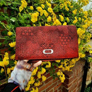 May include: A red clutch with a geometric pattern and a silver clasp. The clutch is rectangular and has a textured surface. The background features yellow flowers and a brick wall.