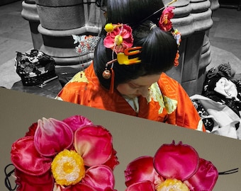 May include: A woman's hair styled with traditional Japanese hair accessories, including red and yellow floral ornaments and hairpins. Two pink and yellow flower hair accessories are in the foreground. The woman is wearing an orange kimono.