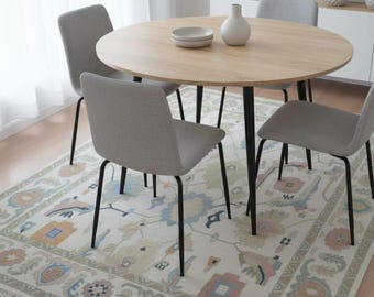 May include: A round wooden dining table with four grey chairs. The table sits on a patterned rug with a cream background and floral and geometric designs. A white vase and two small plates are on the table.