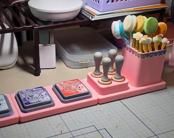 May include: A pink craft organizer holds ink pads, stamps, and brushes. The ink pads are labeled "Distress Oxide." A variety of makeup brushes and blending tools are in a pink container. The organizer sits on a craft table.
