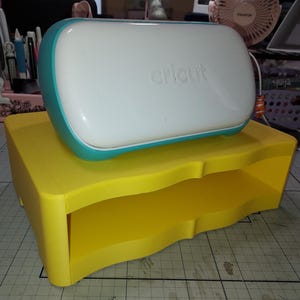May include: A yellow plastic stand with two shelves for a Cricut machine. The Cricut machine is teal and white and has the word "cricut" printed on it.