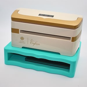 May include: A cream-colored Empress die-cutting machine with gold accents, resting on a turquoise base. The machine has a digital display and the Anna Griffin logo. The base has a wavy design.