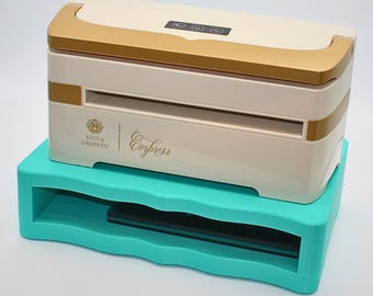 May include: A cream-colored Empress die-cutting machine with gold accents, resting on a turquoise base. The machine has a digital display and the Anna Griffin logo. The base has a wavy design.