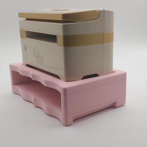May include: A white and gold embossing machine with the text "Embossing Mini" on the front, sitting on a pink plastic stand with a wavy design.
