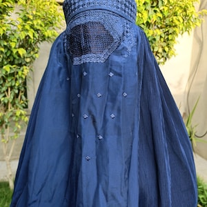 Puede incluir: Una burka azul marino con intrincados detalles bordados. El tocado presenta un patrón geométrico, y la parte trasera tiene un panel decorativo con costuras delicadas. Pequeños acentos bordados están dispersos por la tela.