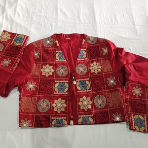 May include: A red jacket with intricate embroidery. The jacket features a V-neck design and long sleeves. The embroidery includes floral and geometric patterns in gold, teal, and white. The jacket is made of a smooth fabric and has a drawstring closure.