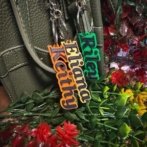 May include: Two personalised keychains with names. One keychain is orange with the name "Keithy" and "Eliana" in black. The other keychain is green with the name "Riley" in black. Both keychains have silver key rings and chains.