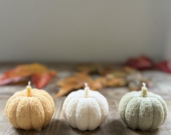 May include: Three pumpkin-shaped candles in shades of orange, white, and green sit on a wooden surface. Each candle has a small wick. The background features blurred fall leaves, creating a seasonal autumn theme.