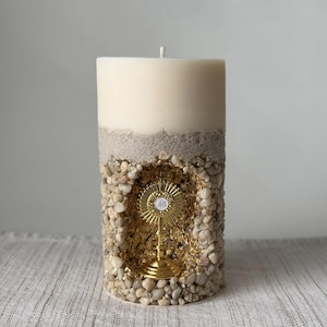 May include: A cream-coloured pillar candle with a decorative design. The candle features a section of small pebbles and a gold-coloured religious symbol. The candle is approximately 15 cm tall.