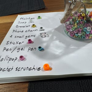 Könnte beinhalten: Ein klares Glasgefäß gefüllt mit bunten Perlen, neben einer Liste von Artikeln, darunter Haargummis, Seife, Armbänder, Handy-Anhänger, Edelsteine, Aufkleber, Stifte, Lutscher und ein gehäkeltes Scrunchie. Die Artikel liegen auf einer weißen Oberfläche.